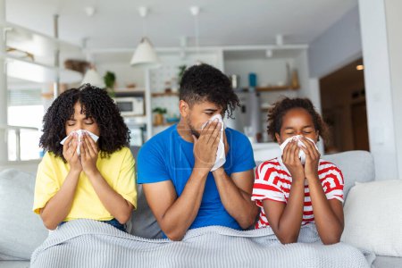 Photo for Flu And Illness Concept. Portrait of sick young African American family blowing noses with napkins together while sitting on the couch covered with blanket. Four unwell people with cold at home - Royalty Free Image