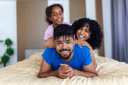 Photo for Portrait of beautiful young mother, father and their daughter looking at camera and smiling while lying on bed leaning on each other - Royalty Free Image