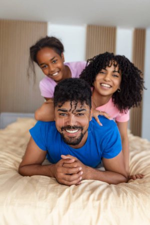 Foto de Retrato de la hermosa madre joven, el padre y su hija mirando a la cámara y sonriendo mientras yacen en la cama apoyados el uno en el otro - Imagen libre de derechos