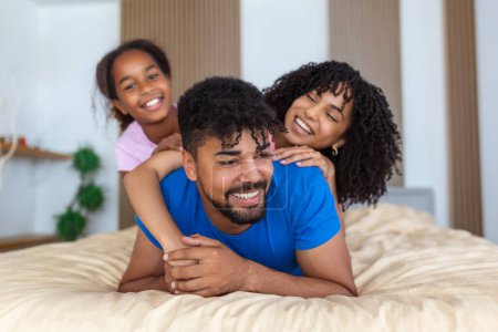 Foto de Sonriente familia afroamericana de tres acostados en la cama uno en la parte superior, uno en el medio y uno en el fondo. - Imagen libre de derechos