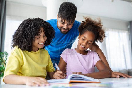 Téléchargez les photos : Vue de face de parents afro-américains aidant leur fille avec des devoirs à table, Photo d'une jeune fille scolarisée à domicile par ses parents - en image libre de droit