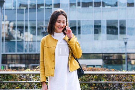 Téléchargez les photos : Asian cheerful young woman walking along the street and calling on phone. Asian woman walking on the street, wearing cute trendy outfit and talking om her smart phone. - en image libre de droit