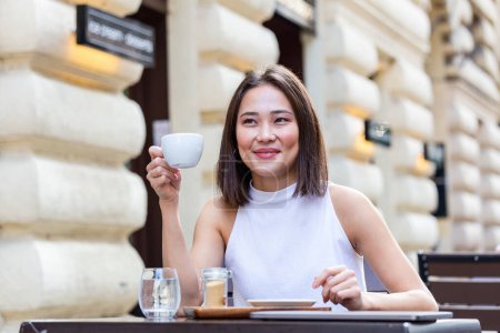 Téléchargez les photos : Beautiful Asian young woman with cup of coffee. Woman enjoys fresh coffee in the morning with sunrise at coffe shop Beautiful woman drinking coffee in the morning sitting outdoors - en image libre de droit