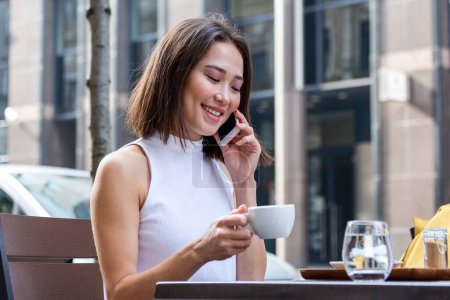 Téléchargez les photos : Smiling Asian woman drinking coffee and using her mobile phone. Satisfied female enjoying cup of coffee. Close up portrait of beautiful girl drinking coffee from a white mug in the coffee shop - en image libre de droit