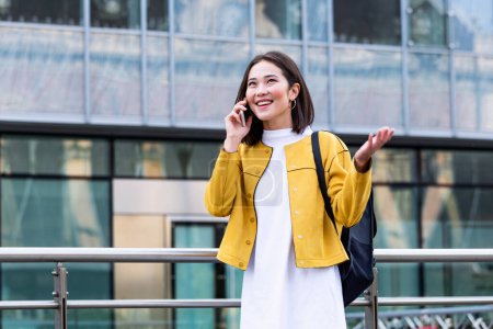 Foto de Asiática alegre joven mujer caminando por la calle y llamando por teléfono. Mujer asiática caminando en la calle, vistiendo lindo traje de moda y hablando om su teléfono inteligente. - Imagen libre de derechos