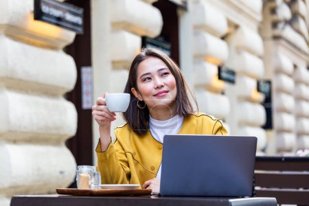 Foto de Young Asian woman sitting in coffee shop at wooden table, drinking coffee and using smartphone.On table is laptop. Girl browsing internet, chatting, blogging. - Imagen libre de derechos