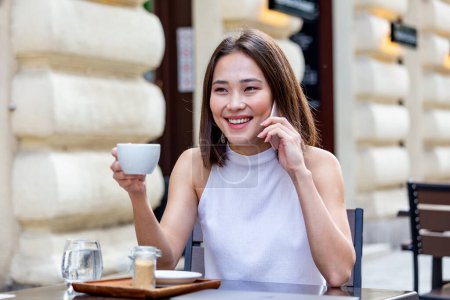 Foto de Smiling Asian woman drinking coffee and using her mobile phone. Satisfied female enjoying cup of coffee. Close up portrait of beautiful girl drinking coffee from a white mug in the coffee shop - Imagen libre de derechos