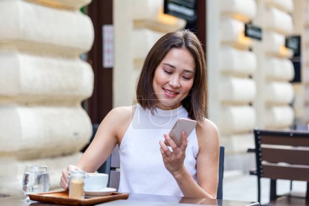 Téléchargez les photos : Smiling Asian woman drinking coffee and using her mobile phone. Satisfied female enjoying cup of coffee. Close up portrait of beautiful girl drinking coffee from a white mug in the coffee shop - en image libre de droit