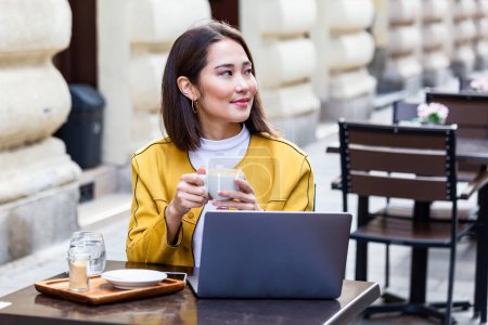 Téléchargez les photos : Jeune femme d'affaires asiatique assise à table et prenant des notes dans un carnet. Sur la table se trouve un ordinateur portable, un smartphone et une tasse de café. Apprentissage étudiant en ligne. Blogueur. - en image libre de droit