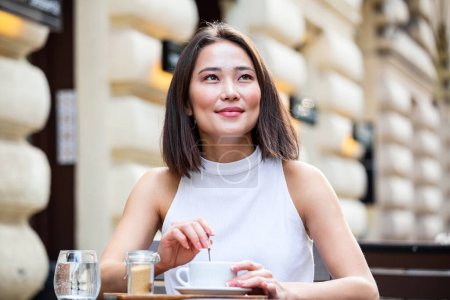 Photo for Young asian business woman sitting at table in cafe shop and writing in notebook. On table is laptop, smartphone and she is holding cup of coffee. Freelancer working in coffee shop. - Royalty Free Image