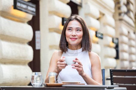 Téléchargez les photos : Femme asiatique souriante buvant du café et utilisant son téléphone portable. Femme satisfaite dégustant une tasse de café. Gros plan portrait de belle fille buvant du café dans une tasse blanche dans le café - en image libre de droit