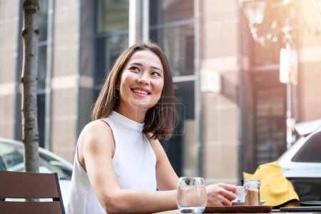 Foto de Beautiful Asian young woman with cup of coffee. Woman enjoys fresh coffee in the morning with sunrise at coffe shop Beautiful woman drinking coffee in the morning sitting outdoors - Imagen libre de derechos