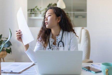 Téléchargez les photos : Doctor professional female Asian doctor wearing uniform taking notes in medical journal, filling documents, patient illness history, looking at laptop screen, student watching webinar - en image libre de droit