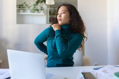 Photo for Portrait of young stressed Asian woman sitting at home office desk in front of laptop, touching aching shoulder with pained expression, suffering from shoulder ache after working on laptop - Royalty Free Image