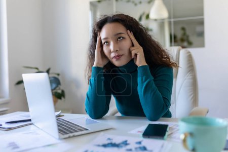 Photo for Exhausted Asian businesswoman having a headache in office. Creative woman working at office desk feeling tired. Stressed casual business woman feeling head pain while overworking on laptop computer. - Royalty Free Image