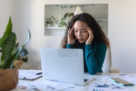 Foto de Exhausted Asian businesswoman having a headache in office. Creative woman working at office desk feeling tired. Stressed casual business woman feeling head pain while overworking on laptop computer. - Imagen libre de derechos