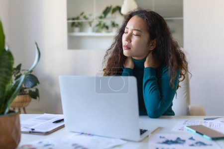 Foto de Feeling exhausted. Frustrated Asian woman looking exhausted and massaging her neck while sitting at her working place - Imagen libre de derechos