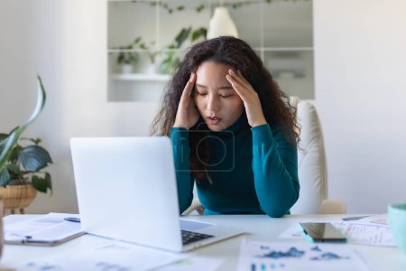 Foto de Exhausted Asian businesswoman having a headache in office. Creative woman working at office desk feeling tired. Stressed casual business woman feeling head pain while overworking on laptop computer. - Imagen libre de derechos