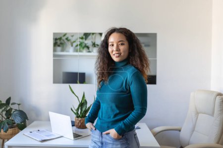 Photo for Smiling young Asian female employee stand at desk in office look in distance thinking or visualizing career success. Happy businesswoman plan or dream at workplace. Business vision concept. - Royalty Free Image