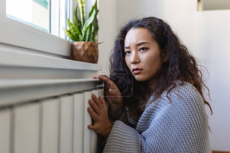 Foto de Unwell Asian woman renter in blanket sit in cold living room hand on old radiator.suffer from lack of heat . Unhealthy young woman struggle from chill freeze at home. No heating concept. - Imagen libre de derechos