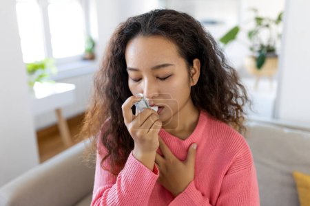 Foto de Asian woman using inhaler while suffering from asthma at home. Young woman using asthma inhaler. Close-up of a young Asian woman using asthma inhaler at home. - Imagen libre de derechos