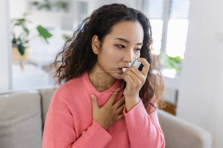 Téléchargez les photos : Young Asian woman using her asthma inhaler on couch at home in the living room - en image libre de droit