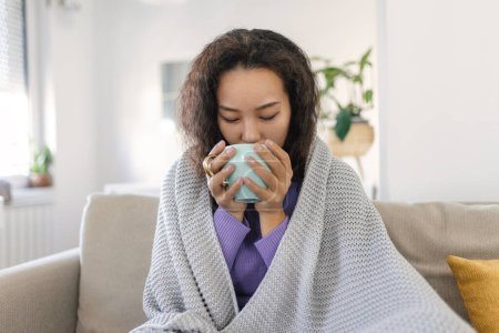 Téléchargez les photos : Sick young Asian woman sitting at home in bed with hot cup of tea and handkerchief. Seasonal colds, cough, runny nose, viral infections, home treatment - en image libre de droit