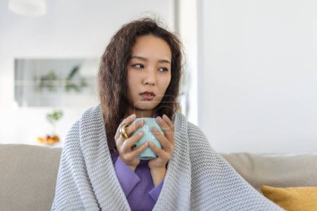 Téléchargez les photos : Sick young Asian woman sitting at home in bed with hot cup of tea and handkerchief. Seasonal colds, cough, runny nose, viral infections, home treatment - en image libre de droit