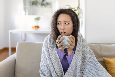 Foto de Sick young Asian woman sitting at home in bed with hot cup of tea and handkerchief. Seasonal colds, cough, runny nose, viral infections, home treatment - Imagen libre de derechos