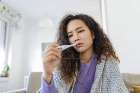 Téléchargez les photos : Asian woman is having flu and she is using thermometer. Sick with a rhinitis womanwith hijab has dripping nose. Woman being sick having flu lying on sofa looking at temperature on thermometer. - en image libre de droit