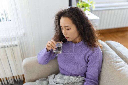 Foto de Young Asian woman holding a glass of water at home. Woman on living room sofa Relaxed While Drinking glass of water. Health benefits of drinking enough water - Imagen libre de derechos