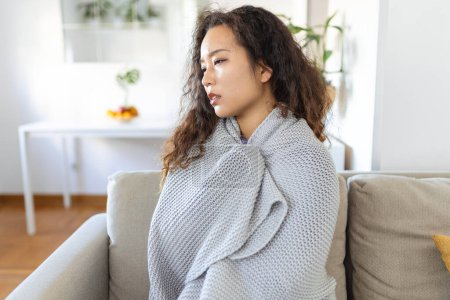 Photo for Sick young Asian woman feeling cold covered with blanket sit on bed, ill black girl shivering freezing warming at home wrapped with plaid, no central heating problem, fever temperature flu concept - Royalty Free Image