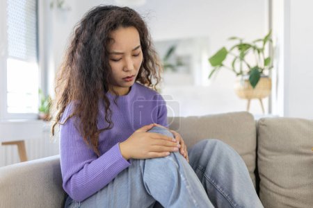 Téléchargez les photos : Young Asian woman is sitting on the couch at home, has pain in the knee joint, holding her leg, osteoarthritis concept. - en image libre de droit
