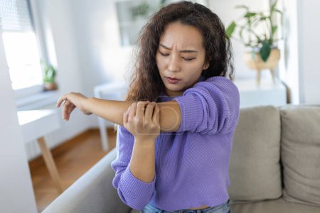 Photo for Unhappy asian woman with elbow pain indoors. office syndrome health care concept. upset frowning asian lady confused looking at arms hurting sitting on couch at home in cozy bright apartment. - Royalty Free Image