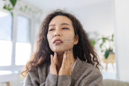 Foto de Close up of young Asian woman rubbing her inflamed tonsils, tonsilitis problem, cropped. Woman with thyroid gland problem, touching her neck, girl has a sore throat - Imagen libre de derechos