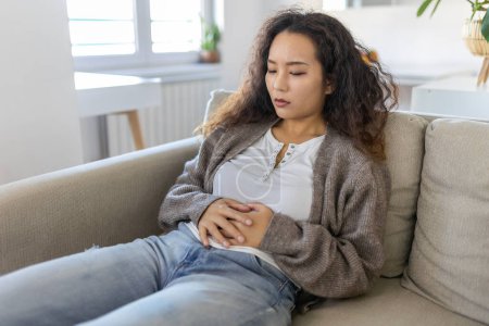 Téléchargez les photos : Asian woman lying on sofa looking sick in the living room. Beautiful young woman lying on bed and holding hands on her stomach. Woman having painful stomachache on bed, Menstrual period - en image libre de droit