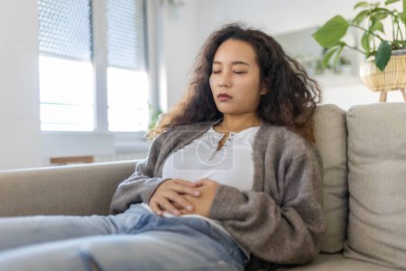 Téléchargez les photos : Asian woman lying on sofa looking sick in the living room. Beautiful young woman lying on bed and holding hands on her stomach. Woman having painful stomachache on bed, Menstrual period - en image libre de droit