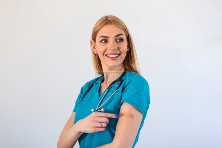 Foto de Portrait of a female doctor smiling after getting a vaccine. Medical worker showing her arm with bandage after receiving vaccination. - Imagen libre de derechos