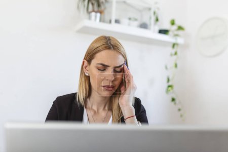Foto de Young frustrated woman working at office desk in front of laptop suffering from chronic daily headaches. Feeling exhausted. Frustrated young woman looking exhausted while sitting at her working place - Imagen libre de derechos