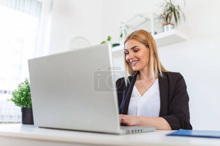 Téléchargez les photos : Young woman working from home, while in quarantine isolation during the Covid-19 health crisis. Portrait of a beautiful young business woman smiling and looking at laptop screen - en image libre de droit