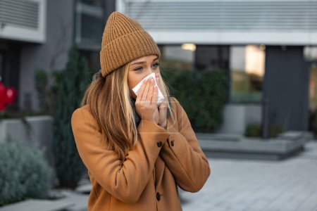 Téléchargez les photos : Ill beautiful young woman sneezing and blowing nose in napkin. Sick businesswoman in business style sneeze at street. Unhealthy employee. Outdoor. Virus symptoms. Cold disease. - en image libre de droit