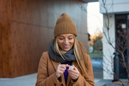 Photo for Positive young female with long blond hair in warm coat applying lip balm while standing on street near building during stroll - Royalty Free Image