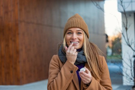 Téléchargez les photos : Jeune femme positive en manteau chaud souriant et regardant loin tout en enduisant de baume à lèvres sur les lèvres pendant la promenade dans la rue de la ville - en image libre de droit