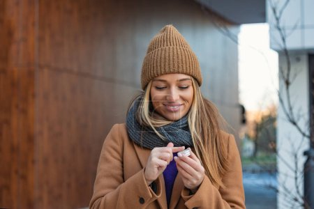 Foto de Positive young female with long blond hair in warm coat applying lip balm while standing on street near building during stroll - Imagen libre de derechos