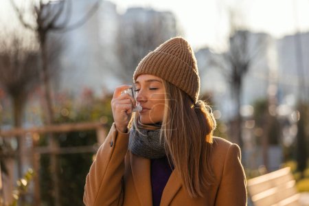 Photo for Side view portrait of an asthmatic woman using a inhaler outdoors in the street. Young woman standing at street, she blocked asthma atack - Royalty Free Image