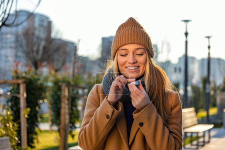 Téléchargez les photos : Positive young female in warm coat smiling and looking away while smearing lip balm on lips during stroll on city street - en image libre de droit
