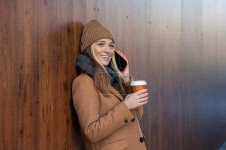 Téléchargez les photos : Joyeux jeune femme portant manteau marchant à l'extérieur, tenant tasse de café à emporter, en utilisant un téléphone mobile - en image libre de droit