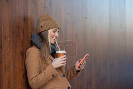 Photo for Cheerful young woman wearing coat using her phone in the sunny city street and drinking take away coffee in paper cup. - Royalty Free Image