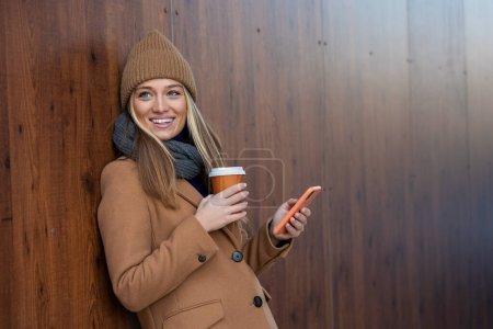 Téléchargez les photos : Cheerful young woman wearing coat using her phone in the sunny city street and drinking take away coffee in paper cup. - en image libre de droit