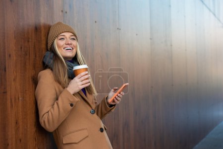 Photo for Cheerful young woman wearing coat using her phone in the sunny city street and drinking take away coffee in paper cup. - Royalty Free Image